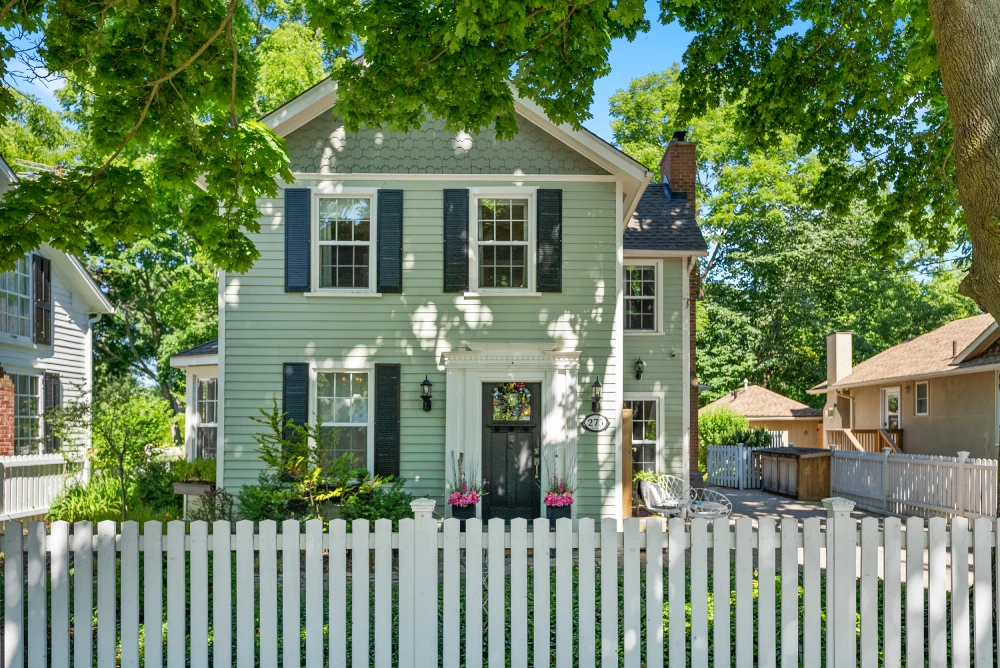 Light green house with white picket fence and leafy trees in front yard