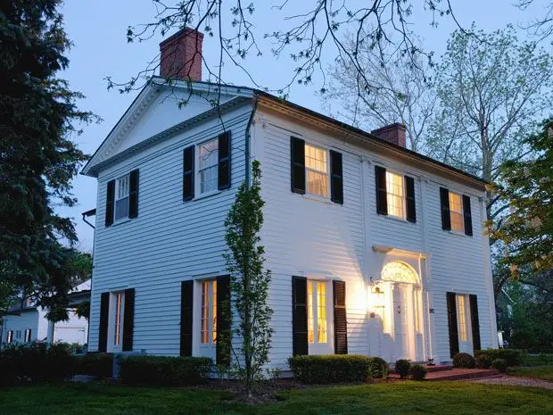 Classic white colonial house at dusk with black shutters and warm interior lights
